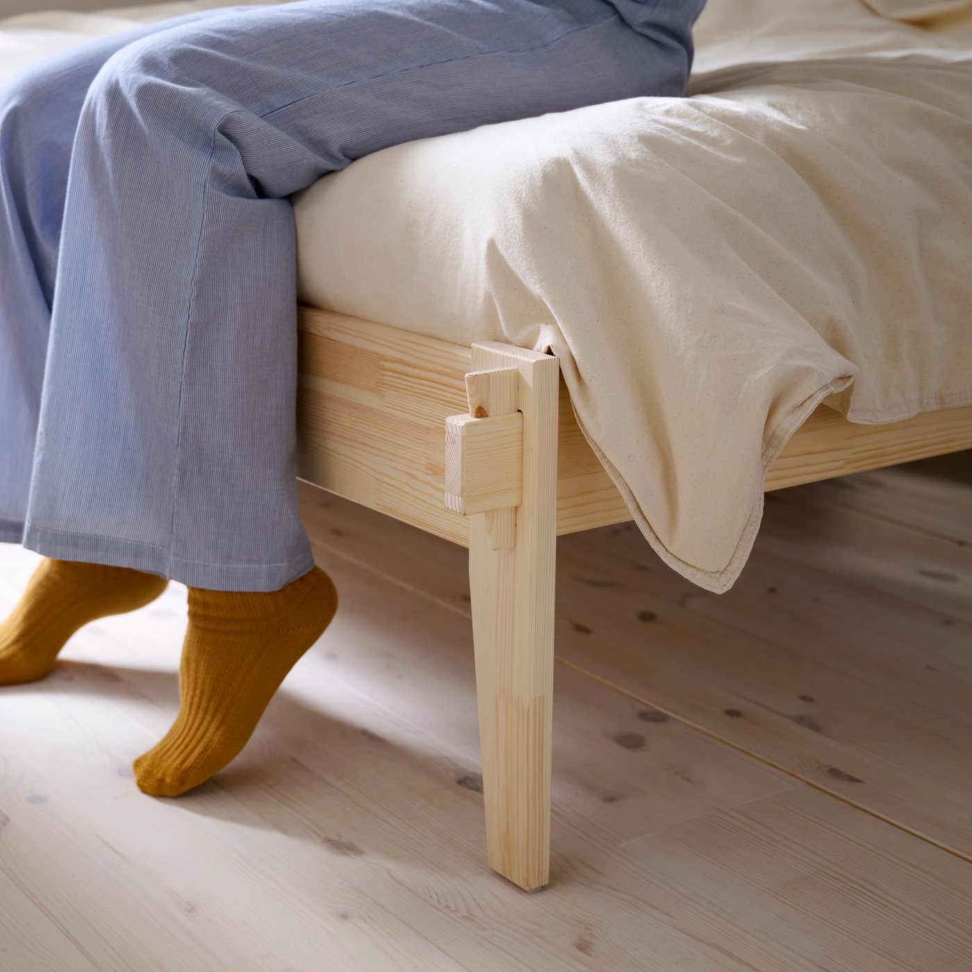 A close up of someone’s legs in blue pyjamas sitting on the edge of a wooden TAXÅSKLINT bed featuring unique joinery.