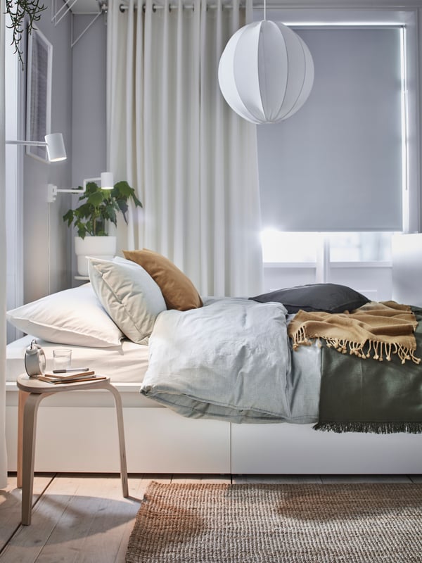 A white MALM bed with 2 storage boxes covered with various linen, in front of a window with MERETE room-darkening curtains.