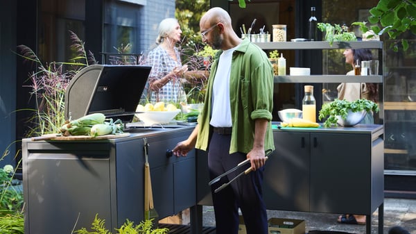 A man is in a garden and tends the barbecue in the BÅTSKÄR outdoor kitchen while two women chat in the background.