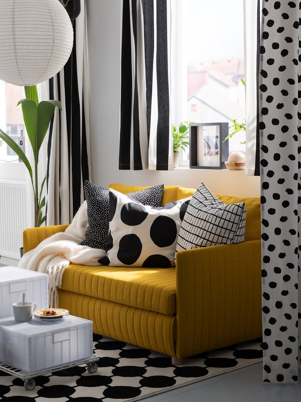 Vorhänge aus schwarz-weiß gestreiftem VARGTÖREL-Zuschnittstoff hängen in einem Raum neben einem gelben FRIDHULT-Bettsofa. Darauf liegen Sofakissen im Skandi Stil und eine Wolldecke.