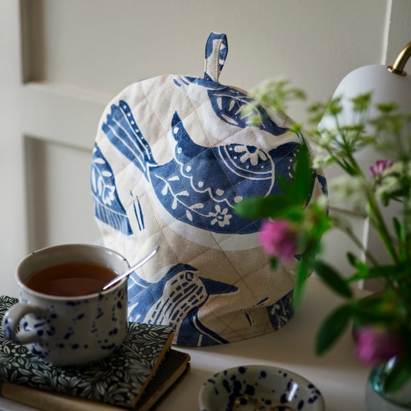 A tea cosy crafted from beige and blue BITTERSÖTA pre-cut fabric sits atop a teapot, next to a mug and some notebooks.