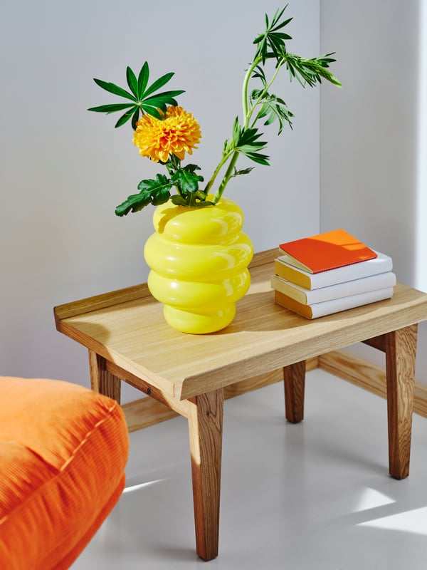 Eine knallig gelbe Vase mit bauchigen Wellen, steht auf einem Beistelltisch, daneben ein Stapel Bücher, darin eine Blüte und zwei Zweige grüne Blätter.
