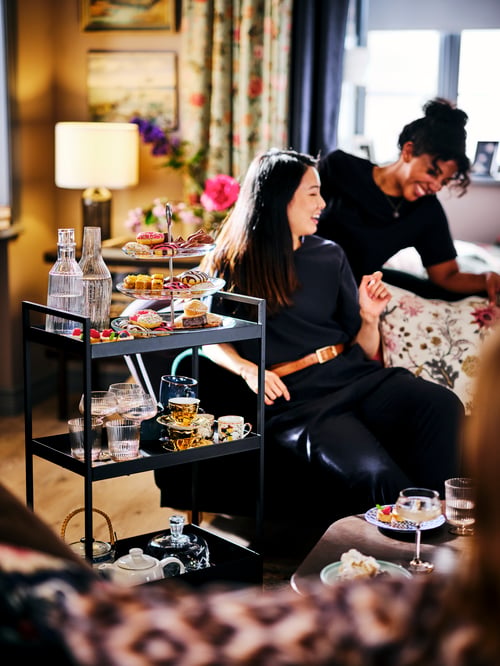 Deux femmes qui rient assises à côté d’une desserte NISSAFORS noire avec des friandises, des verres, des théières et des tasses de thé.