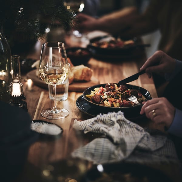Hands holding a knife and a fork chop a bowl of pasta and meatballs. Beside the bowl is a napkin and a wine glass.