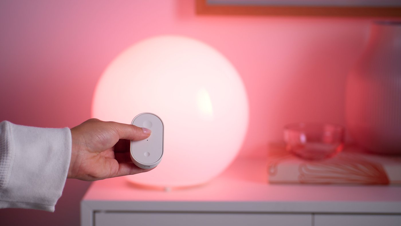 Hand holding a white remote control beside a round smart table lamp glowing warm pink on a white cabinet.