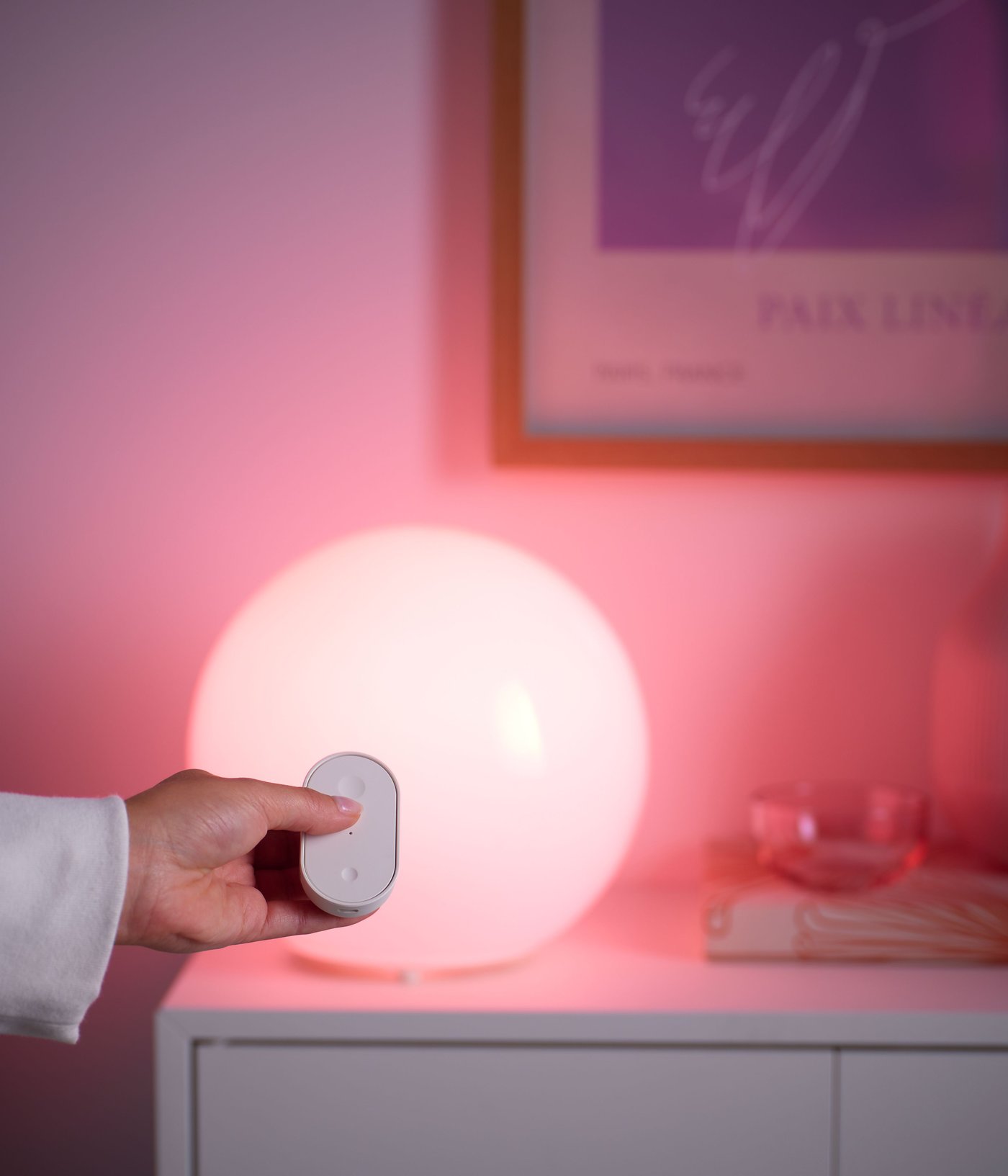 Hand håller en vit fjärrkontroll framför en rund smart bordslampa med varmt rosa sken på en vit byrå.