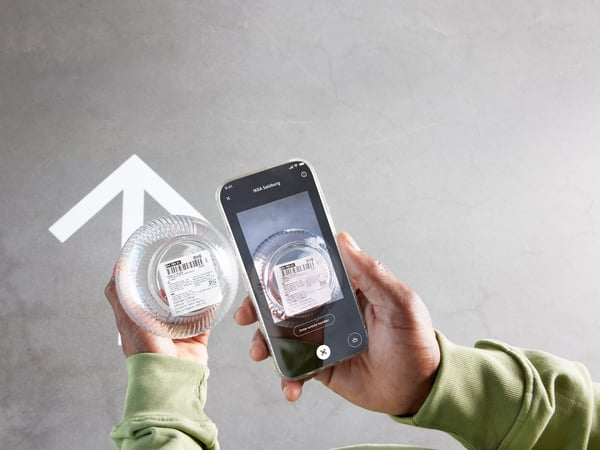 A person holds a smartphone scanning a barcode on a product, above a large white arrow on a gray floor.