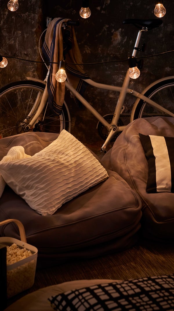 Dettaglio di alcune poltrone da terra con cuscini. Dietro, una bicicletta e alcune lucine accese che illuminano la sera.