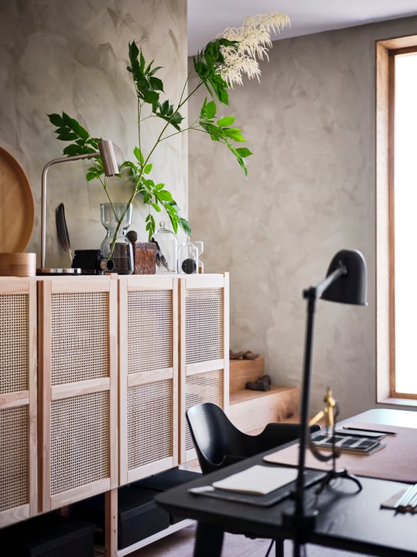 A room with window and lattice-fronted wall storage unit with plant and accessories on it and black desk, chair and lamp.