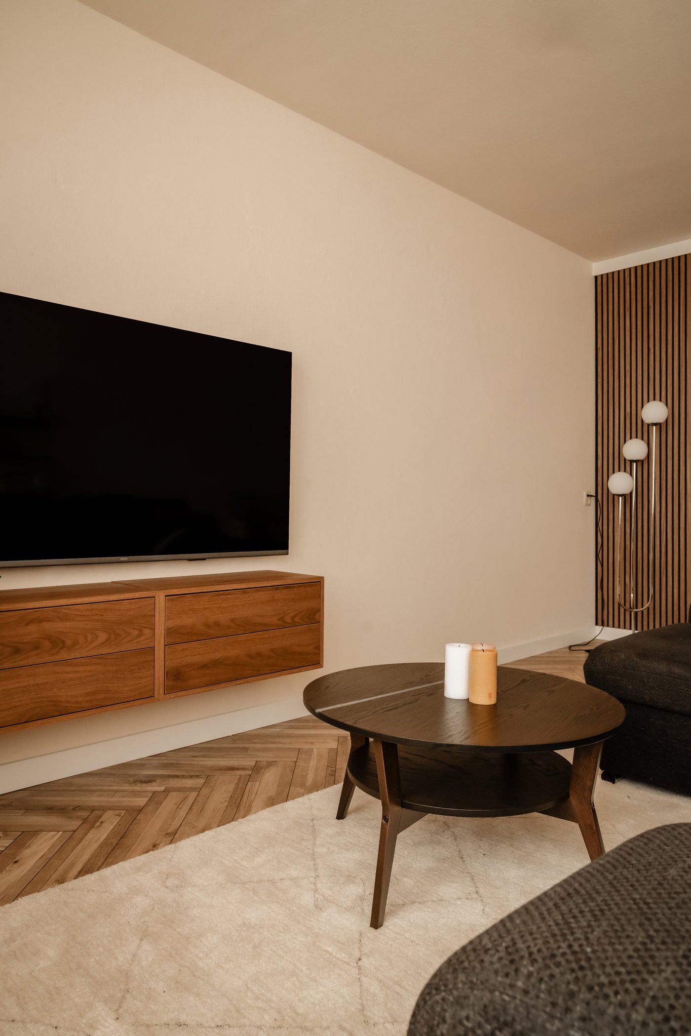 Woonkamer met TV aan de muur, waaronder twee EKET kastjes in bruin walnootpatroon hangen. Verder is een deel van de donkere bank zichtbaar, waaronder een TVERSTED beige vloerkleed ligt met daarop een ronde JAKOBSFORS salontafel in eiken fineer. In de achtergrond staat een SIMRISHAMN staande lamp van glas en chroom.