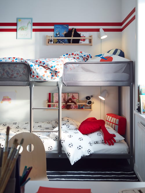 The head of a TUFFING bunk bed in a white room with a red stripe on the wall. A shelf with books is on the wall by each bed.
