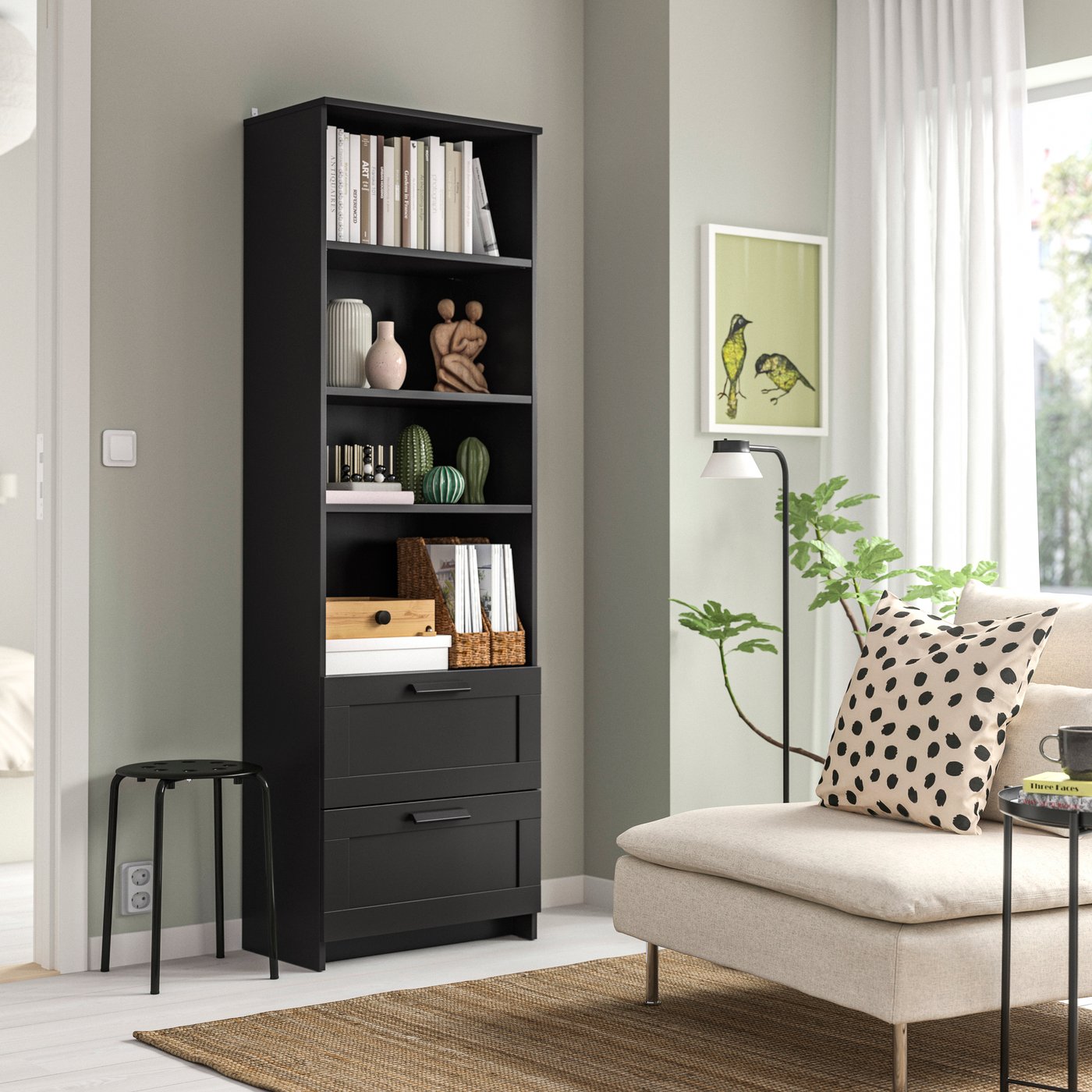 Schwarzes BRIMNES Bücherregal mit Büchern und Wohnaccessoires neben einem Sofa in einem hellen Wohnzimmer.