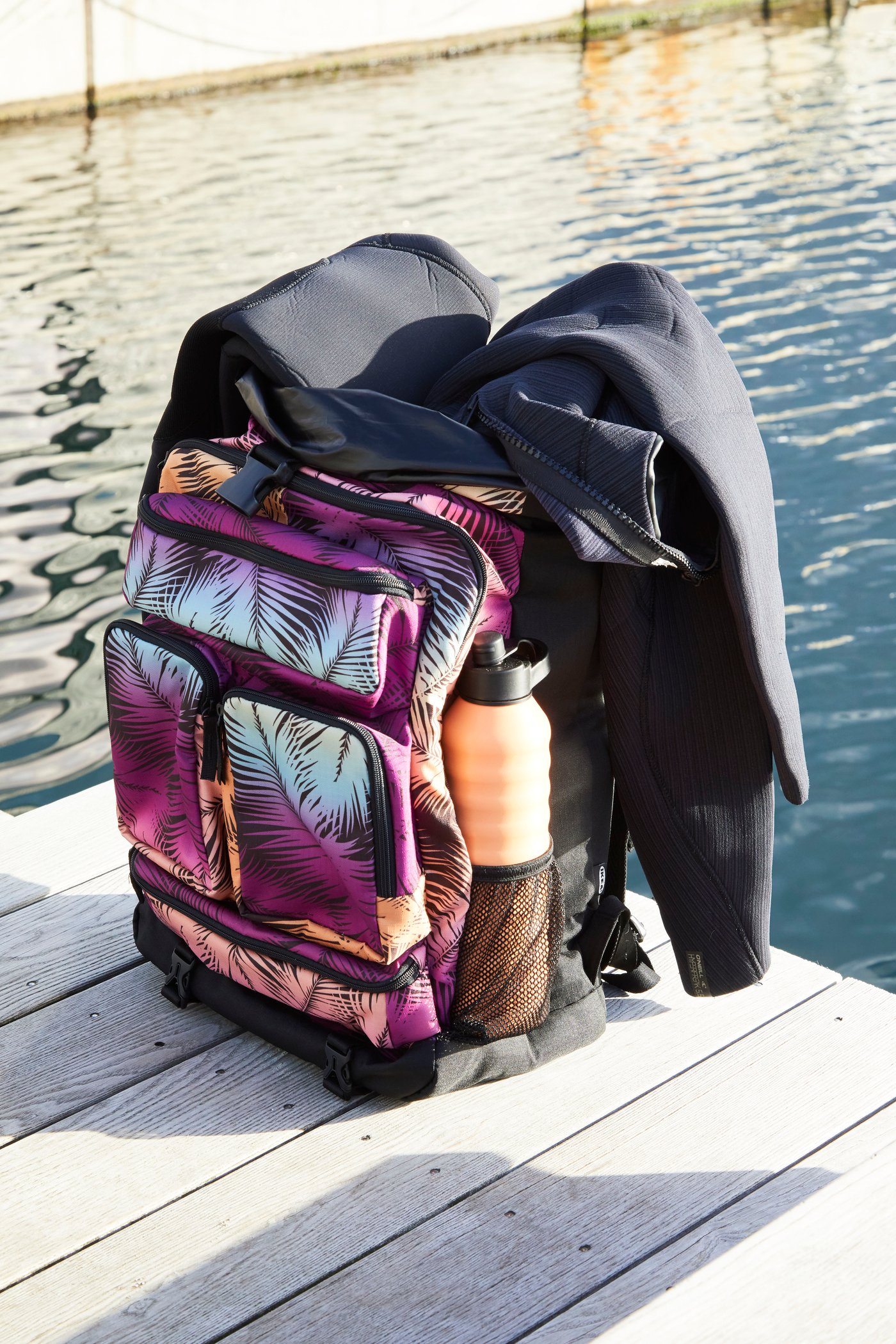 KÅSEBERGA beach towel in a colourful palm-inspired print hanging over the shoulders of a man holding a yellow surfboard.