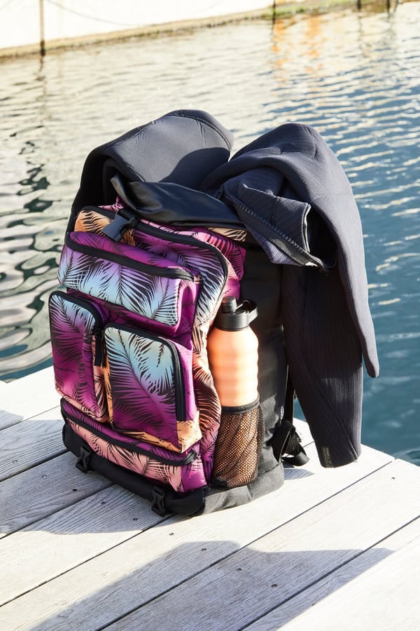 KÅSEBERGA beach towel in a colourful palm-inspired print hanging over the shoulders of a man holding a yellow surfboard.