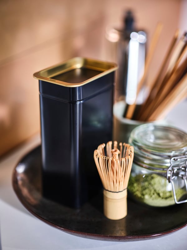 A brown dish on a surface, holding a black BLOMNING coffee/tea tin, a glass jar of matcha and a matcha whisk.