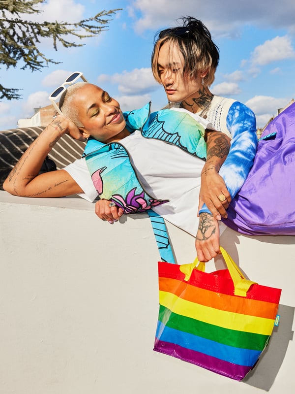 Two people out in the sun, laying on a concrete bench while holding a small STORSTOMMA bag, the IKEA Pride bag. 