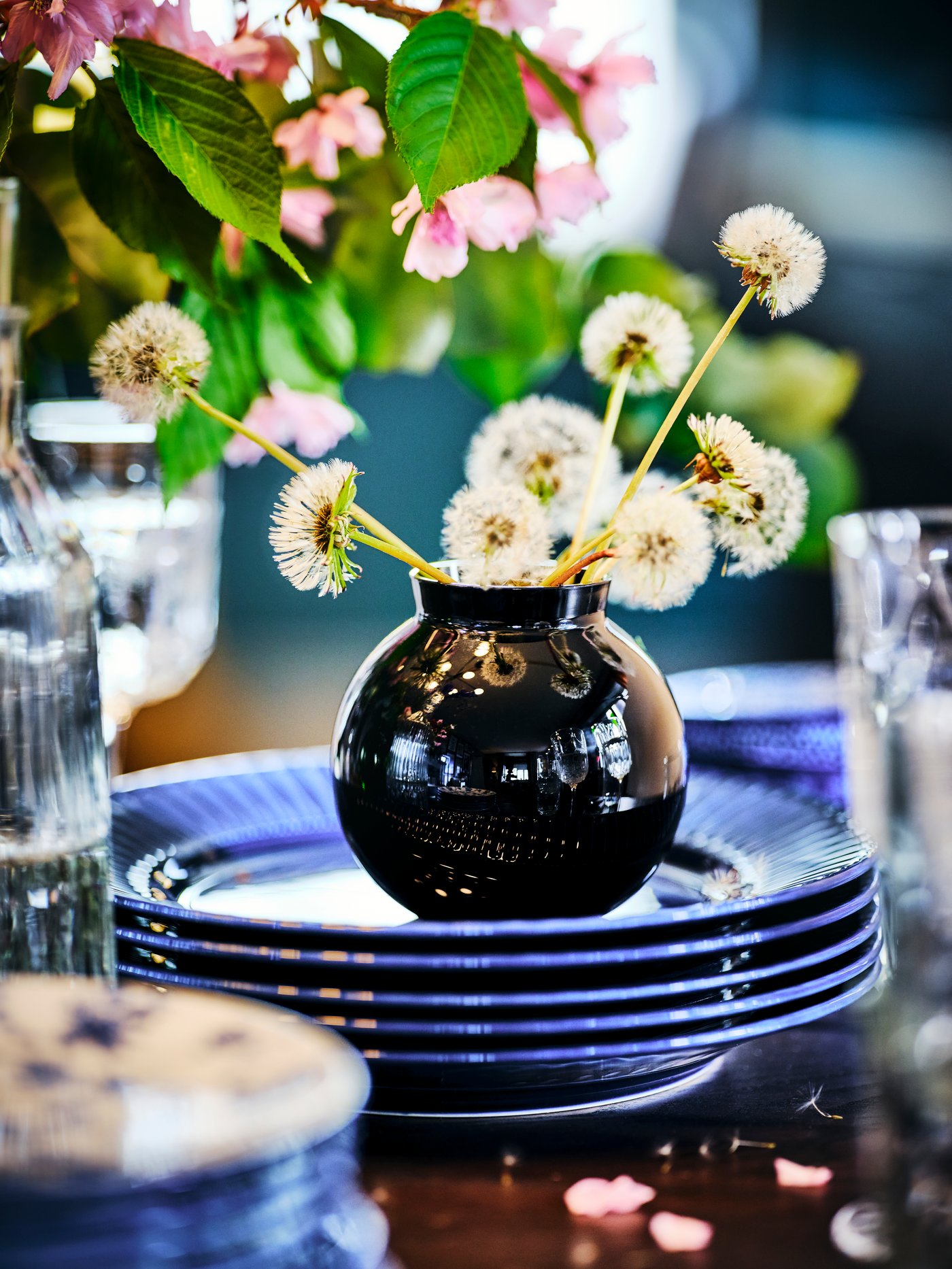 A black KONSTFULL vase of dandelions atop five stacked blue earthenware STRIMMIG plates on a wooden surface.