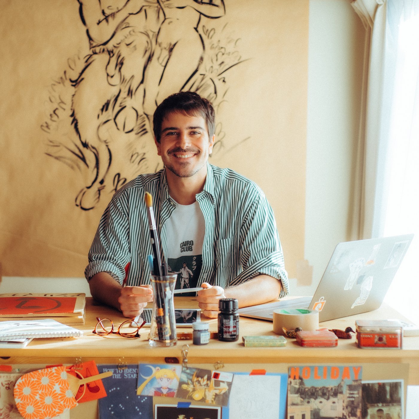 A smiling man sitting in his studio. There is drawing behind him on the wall. He is behind his desk full of art supplies.