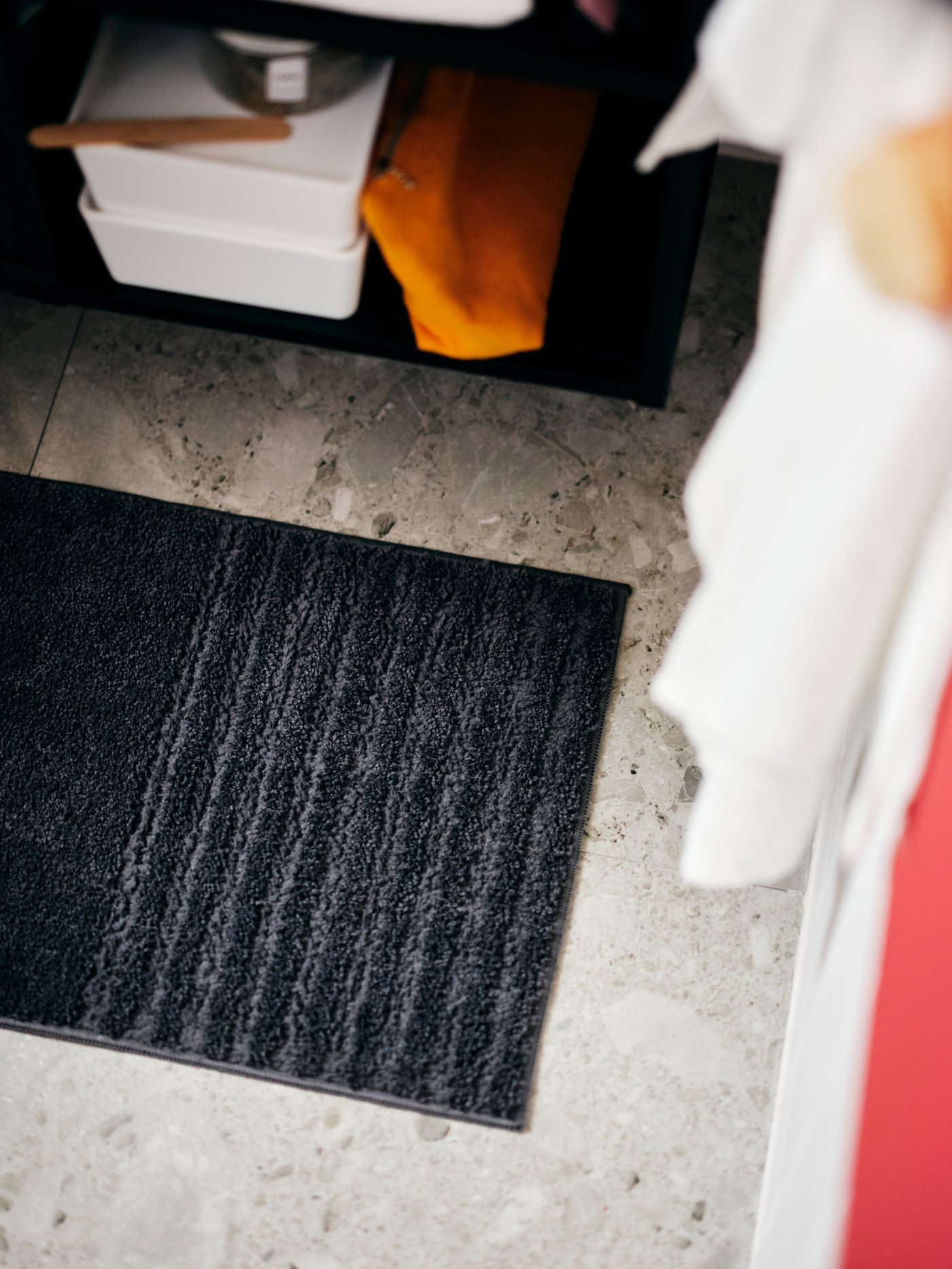 A bathroom corner with hanging towels, accessories on a shelf and a dark-grey VINNFAR bathroom mat on the tiled floor.