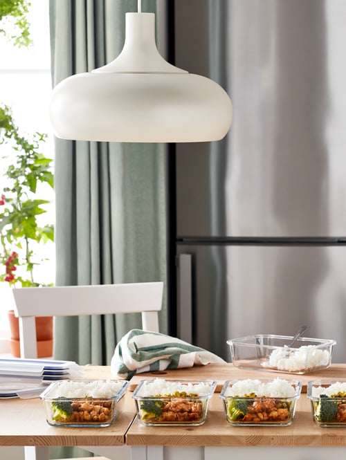 Dining table with white pendant lamp above it, multiple food containers with meal prep components, fridge in the background.