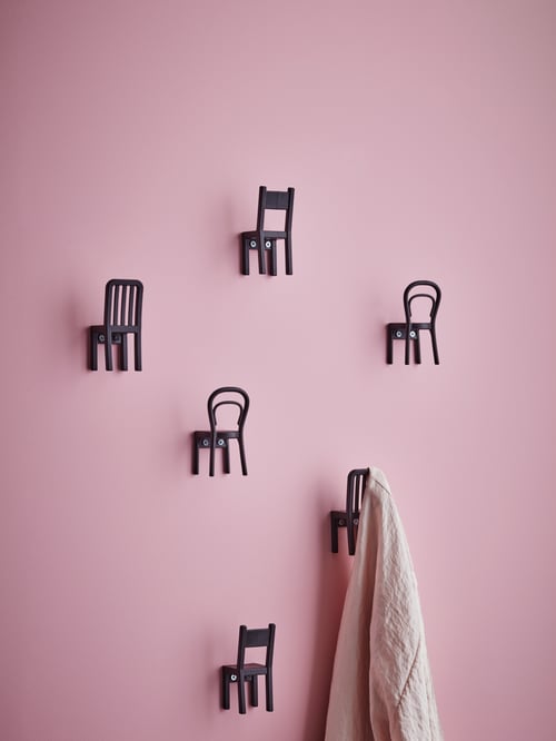 Pink wall with six plastic hooks shaped like different types of chairs, one of them holding a white scarf.