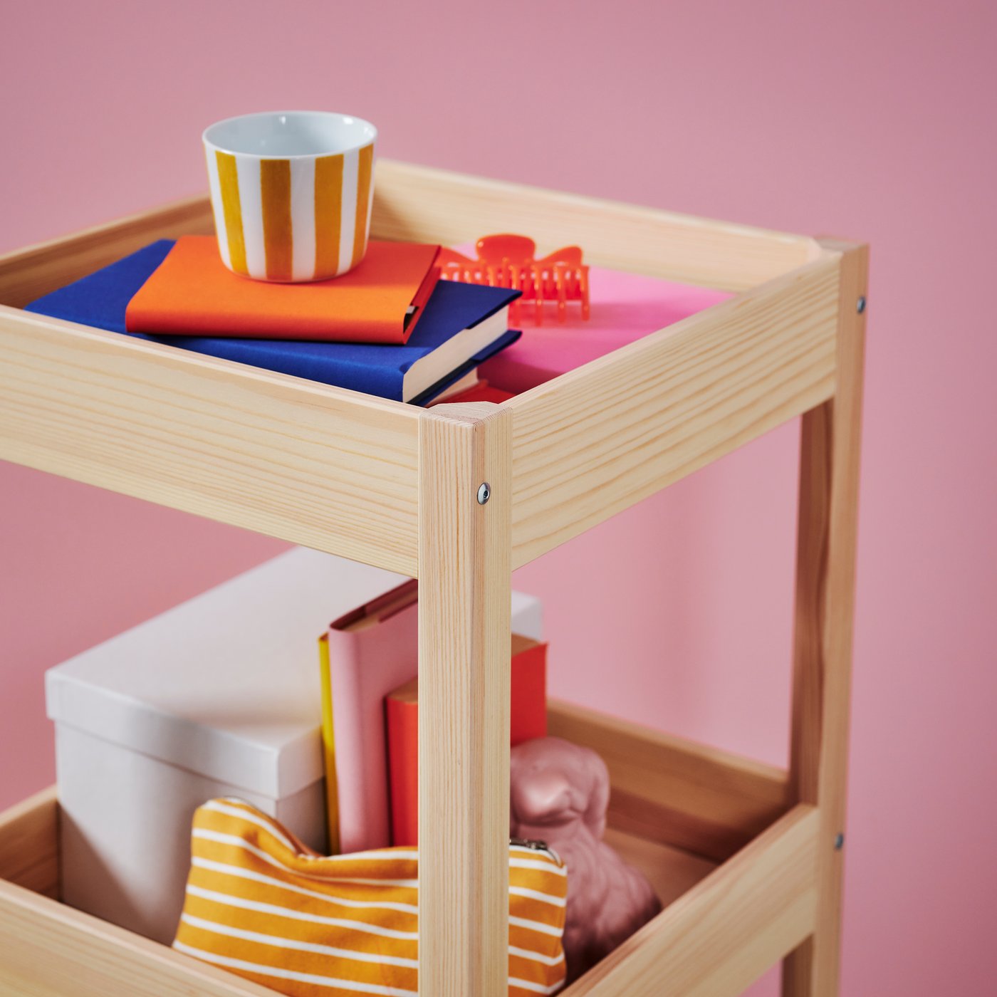 Close-up of colourful cup, books and other objects on light wood trolley with pink background.
