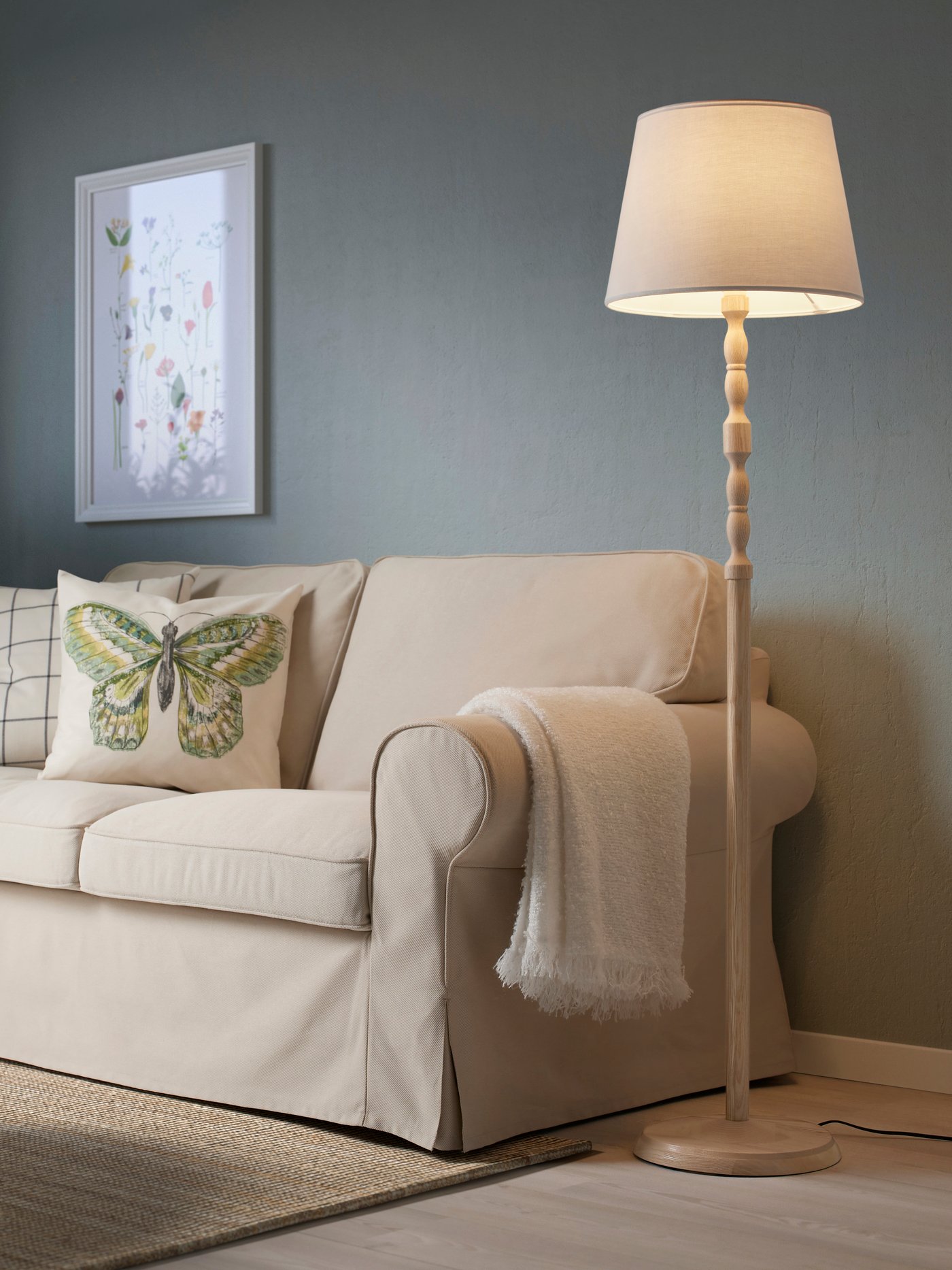 Een landelijke woonkamer met een beige zitbank en een houten KINNAHULT vloerlamp.