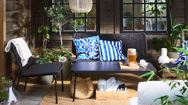 A cosy terrace with black TEGELÖN lounge furniture, blue and white pillows, and decorative SOLVINDEN lighting.