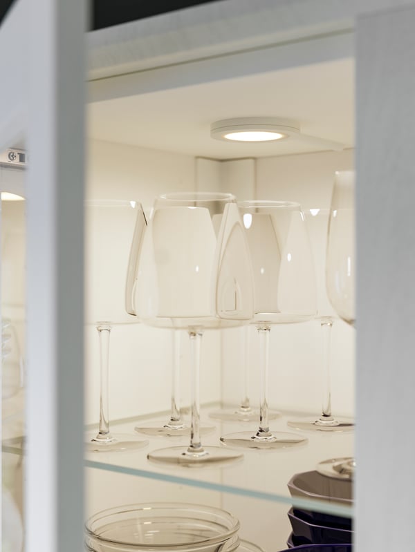 Several glasses are standing on a shelf in a cabinet with white ENKÖPING glass fronts, lit up by a round MITTLED spotlight.