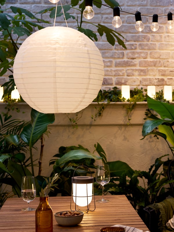 A cosy patio in evening light with an acacia table, SOLVINDEN LED pendant and table lamps, and plenty of large, green plants.