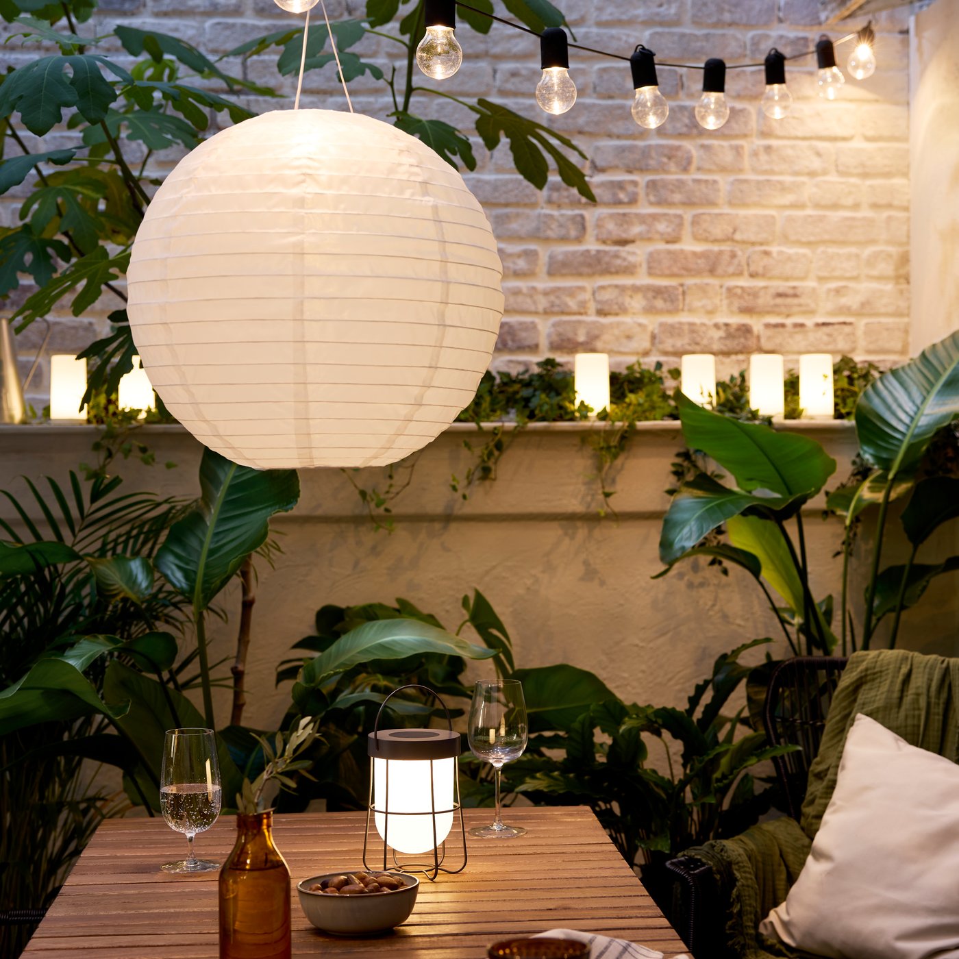 A cosy patio in evening light with an acacia table, SOLVINDEN LED pendant and table lamps, and plenty of large, green plants.