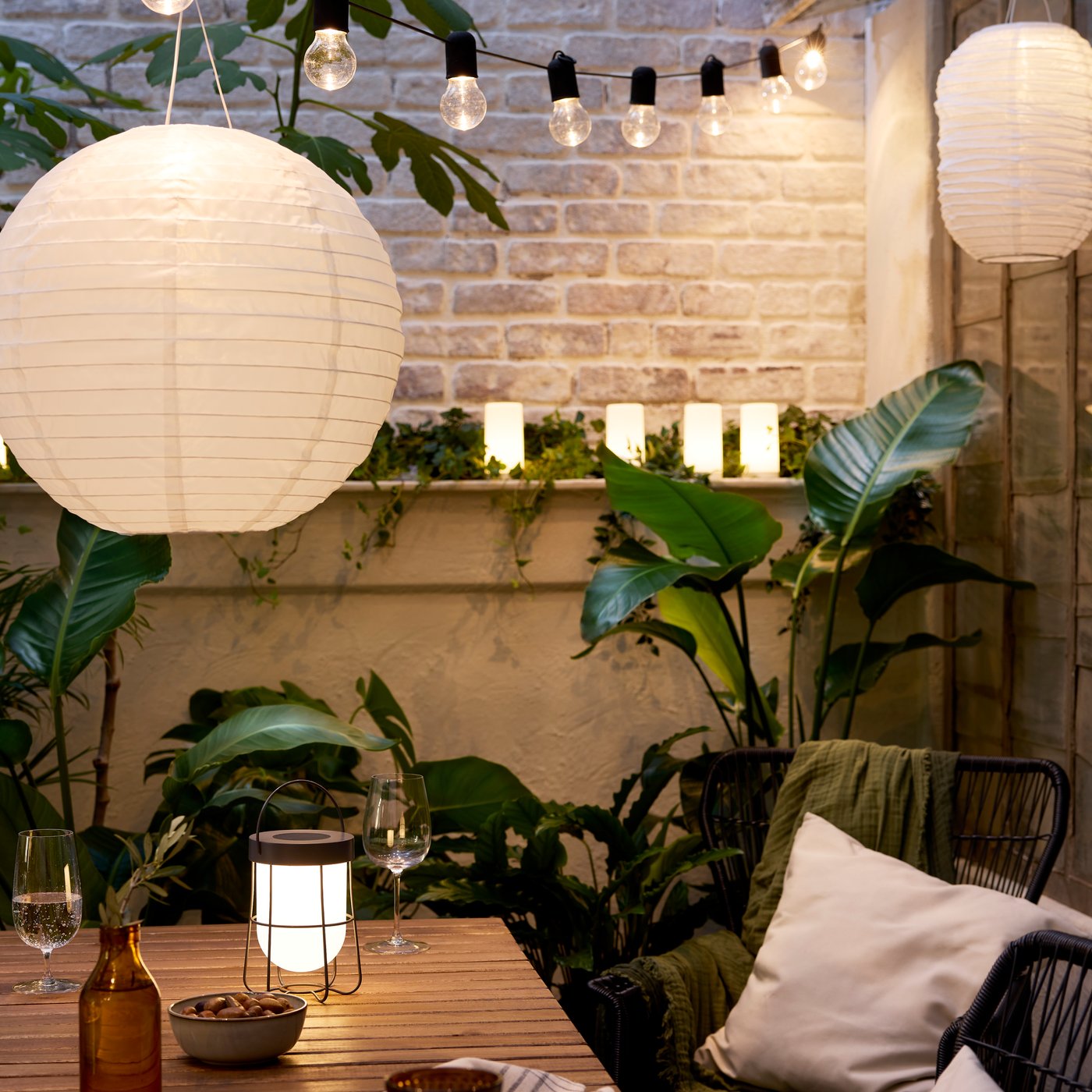 Nella luce della sera, un accogliente patio con un tavolo in acacia, lampade a LED a sospensione REGOLIT / HEMMA e da tavolo SOLVINDEN e tante piante verdi grandi.