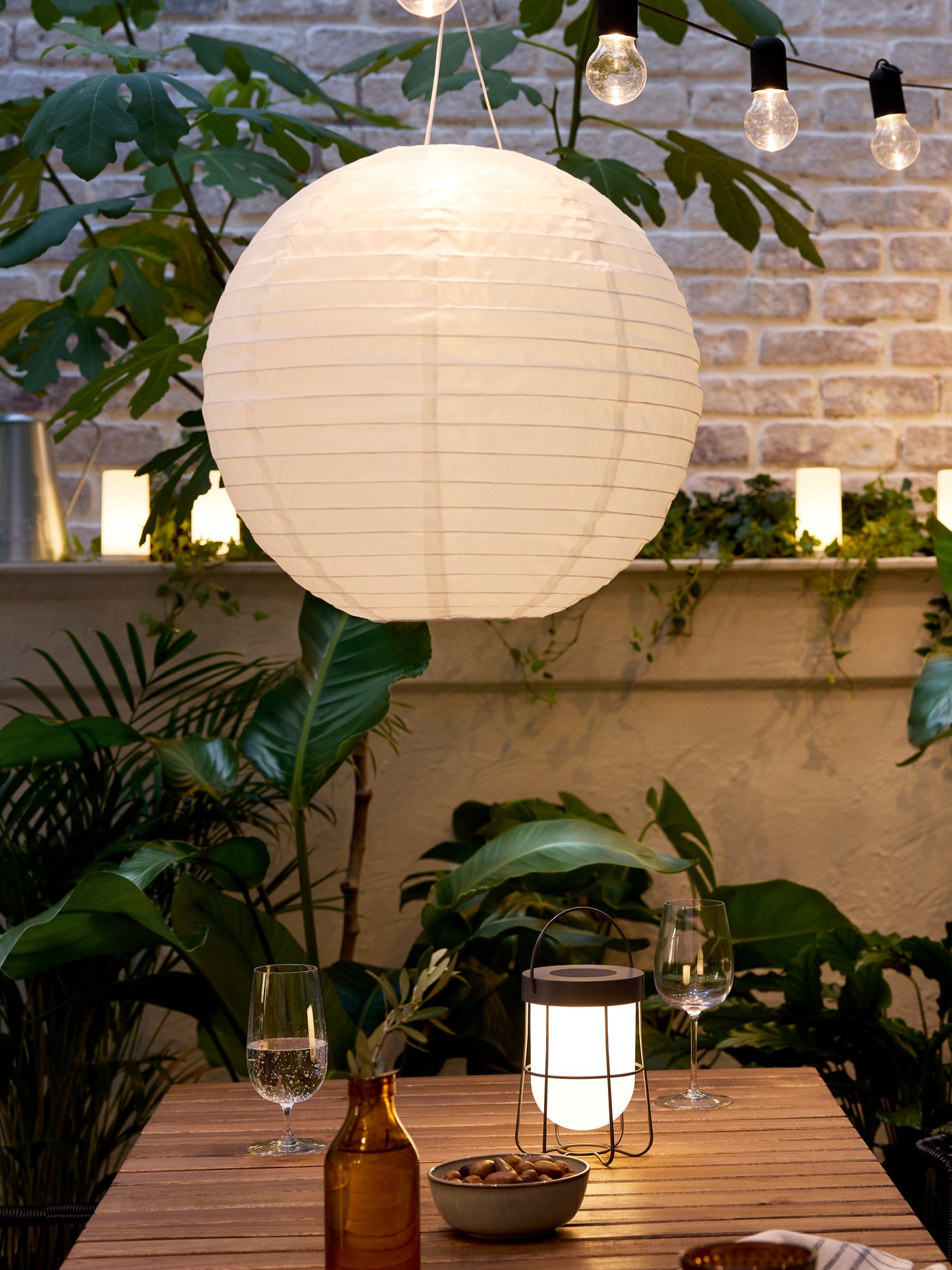 A cosy patio in evening light with an acacia table, SOLVINDEN LED pendant and table lamps, and plenty of large, green plants.