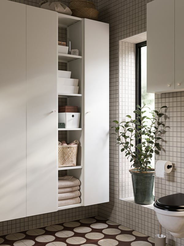 Casa de banho com armários altos ENHET com quatro prateleiras e portas ENHET em branco e caixas KUGGIS com tampas em branco.