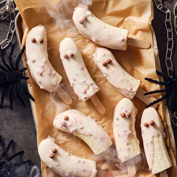 Frosty banana ghost popsicles in BELÖNING white chocolate with raspberries on a baking sheet with toy spiders around them.