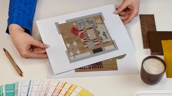 Projektantka wnętrz IKEA prezentująca kartki z wydrukiem rzutu z góry zaprojektowanego pomieszczenia. Obok na blacie leży wzornik kolorów i próbki materiałów.