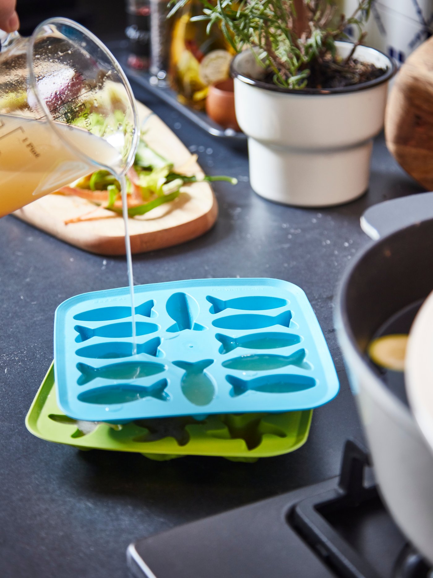 PLASTIS isterningbakker på et køkkenbord. Den øverste er ved at blive fyldt med grøntsagsbouillon, der hældes ud af en glaskande.