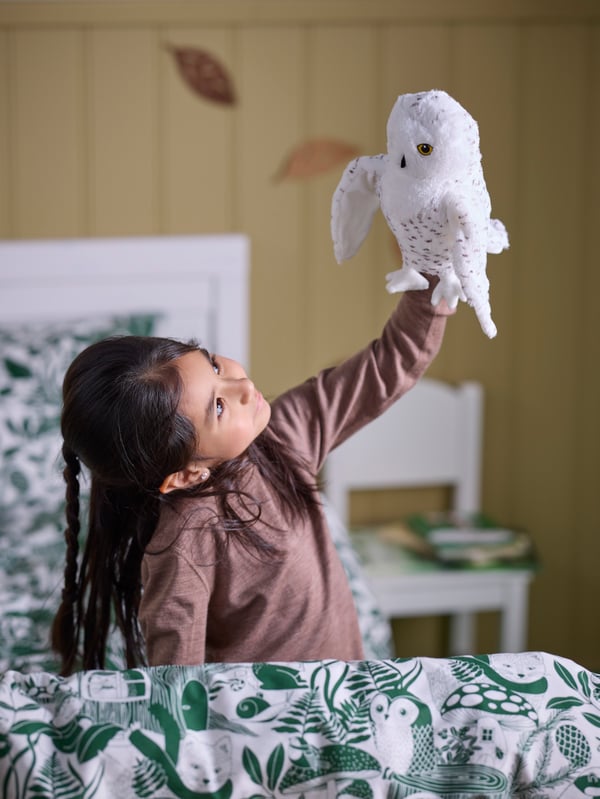 Una niña juega con un peluche de búho SKOGSDUVA en una cama con ropa de cama SKOGSDUVA con estampado de bosques.