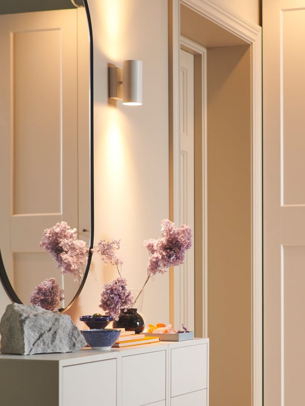 A NYMÅNE wall up/downlighter is mounted on a wall above some flowers in a vase on a cabinet and next to a LINDBYN mirror.