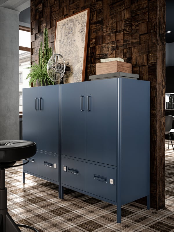 Two dark blue cabinets with doors and drawers with boxes, a framed old map, plants and a retro electric fan on top.