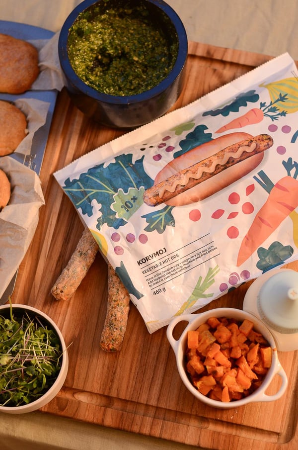 Un tagliere di legno con grissini, un colorato involucro per sandwich, pesto, microverdure, patate dolci a cubetti e una bottiglia di salsa bianca che ricordano un picnic rustico.