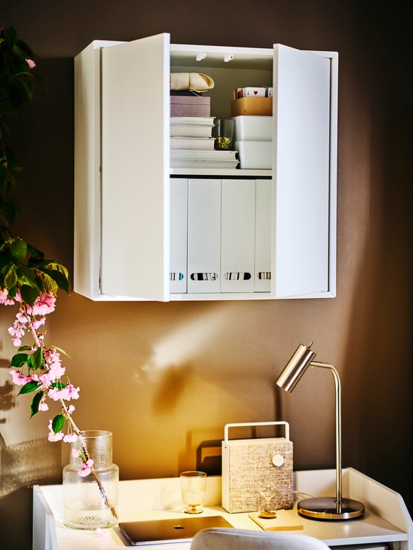 A white EKET cabinet with two doors partially open revealing books inside, above a white HAUGA desk with a VIRRMO work lamp.