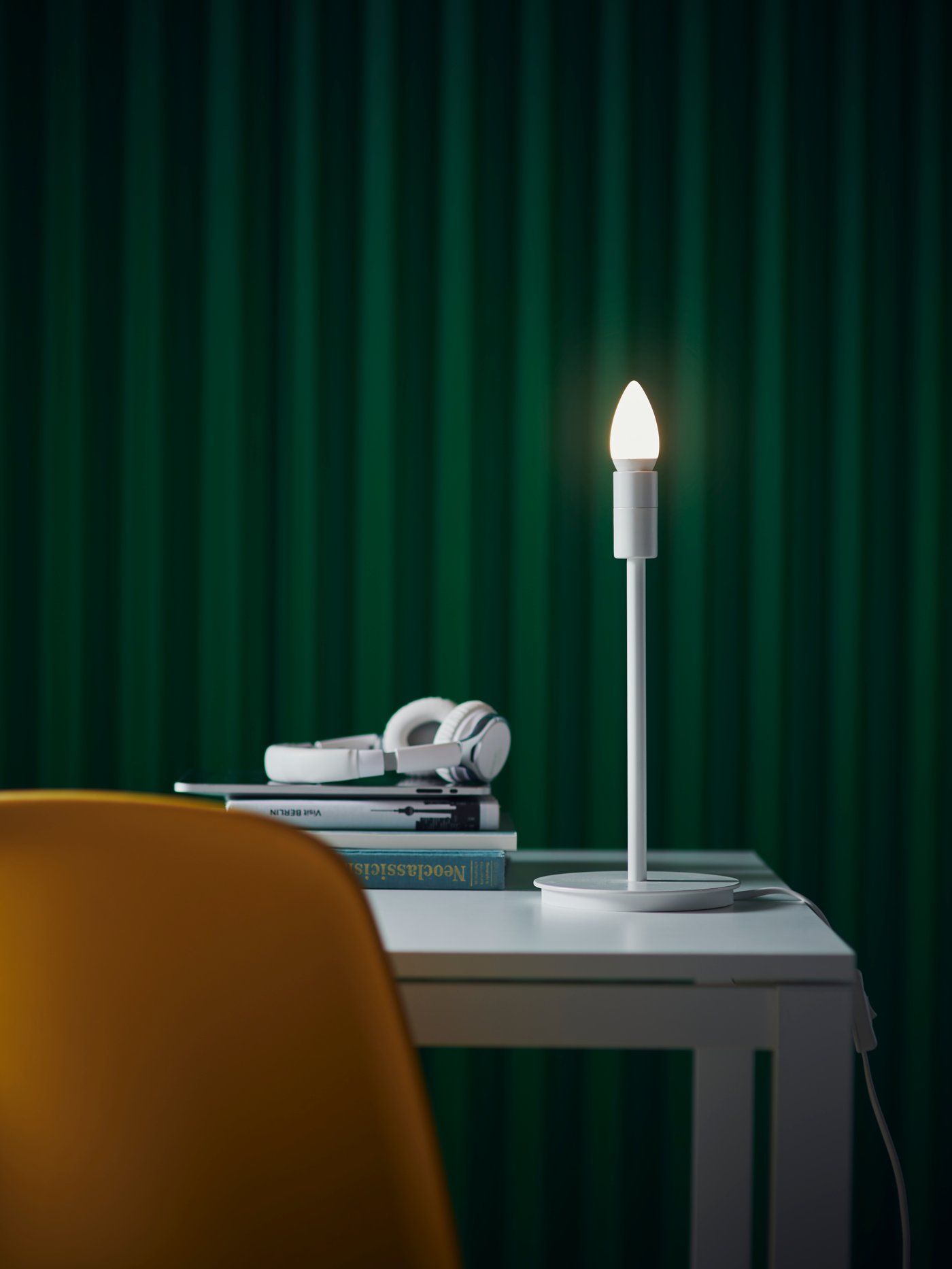 Een witte bureaulamp schijnt op een witte tafel met een stapel boeken en een witte koptelefoon. Op de voorgrond staat een oranje stoel en de muur erachter is donkergroen.