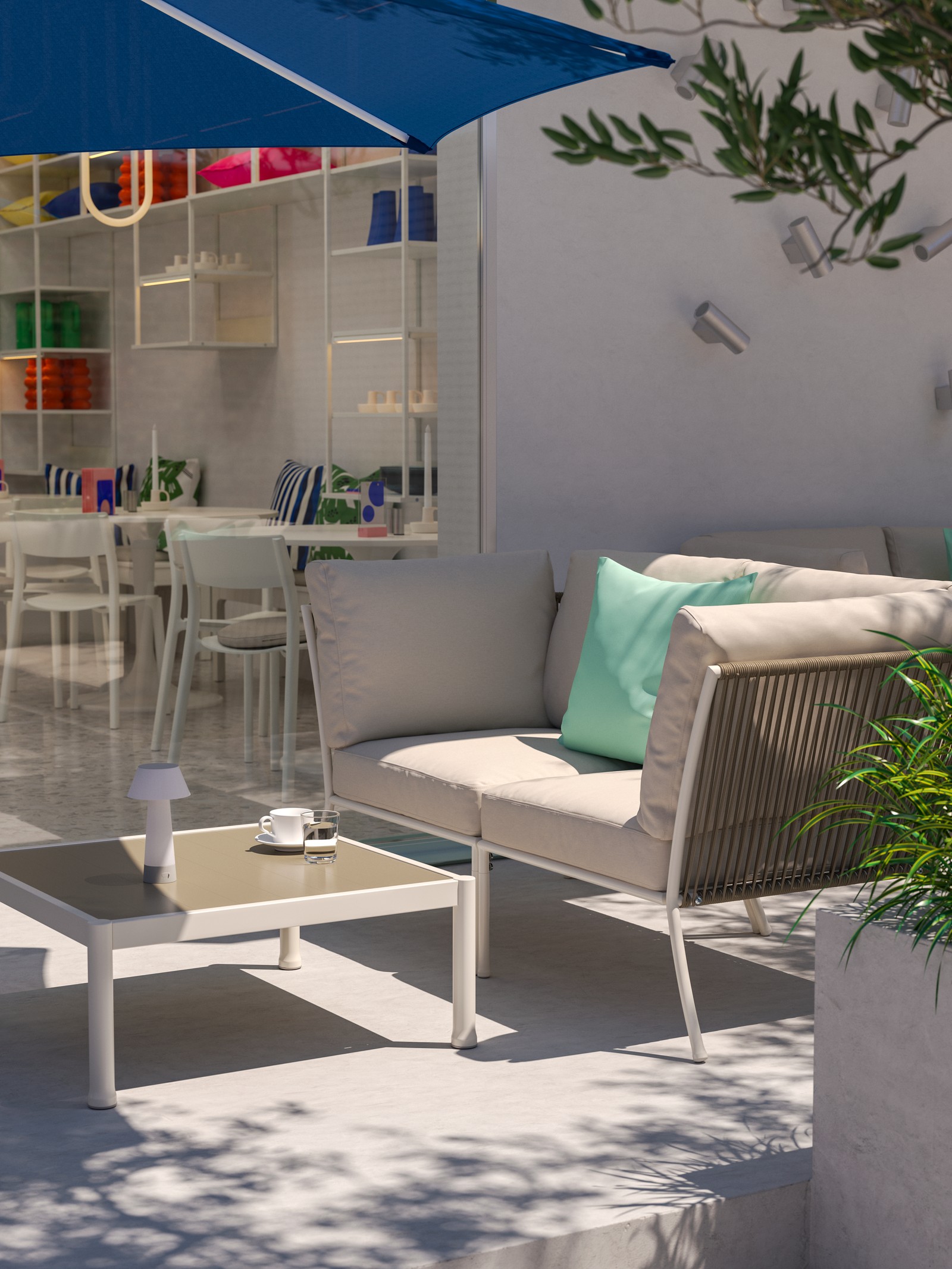 A sunny outdoor café lounging area with a 2-seat white/beige SEGERÖN outdoor conversation set, a blue parasol and greenery.