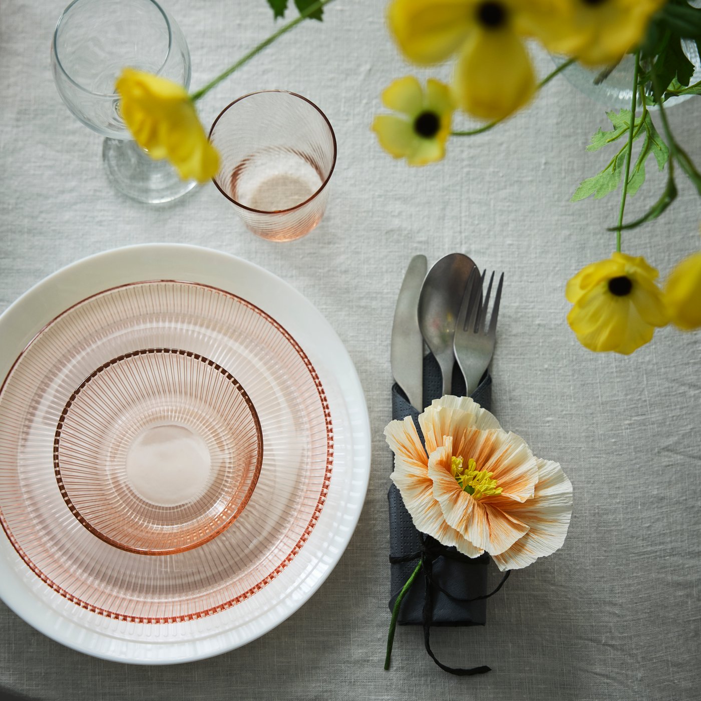 A pink HAVSGÄDDA bowl with a stripe pattern is set on a table with a tablecloth, with cutlery, glasses and flowers.