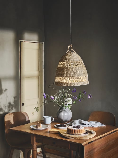 A beige pendant lamp shade hangs above a rectangular drop leaf table with tableware items, and brown chairs.
