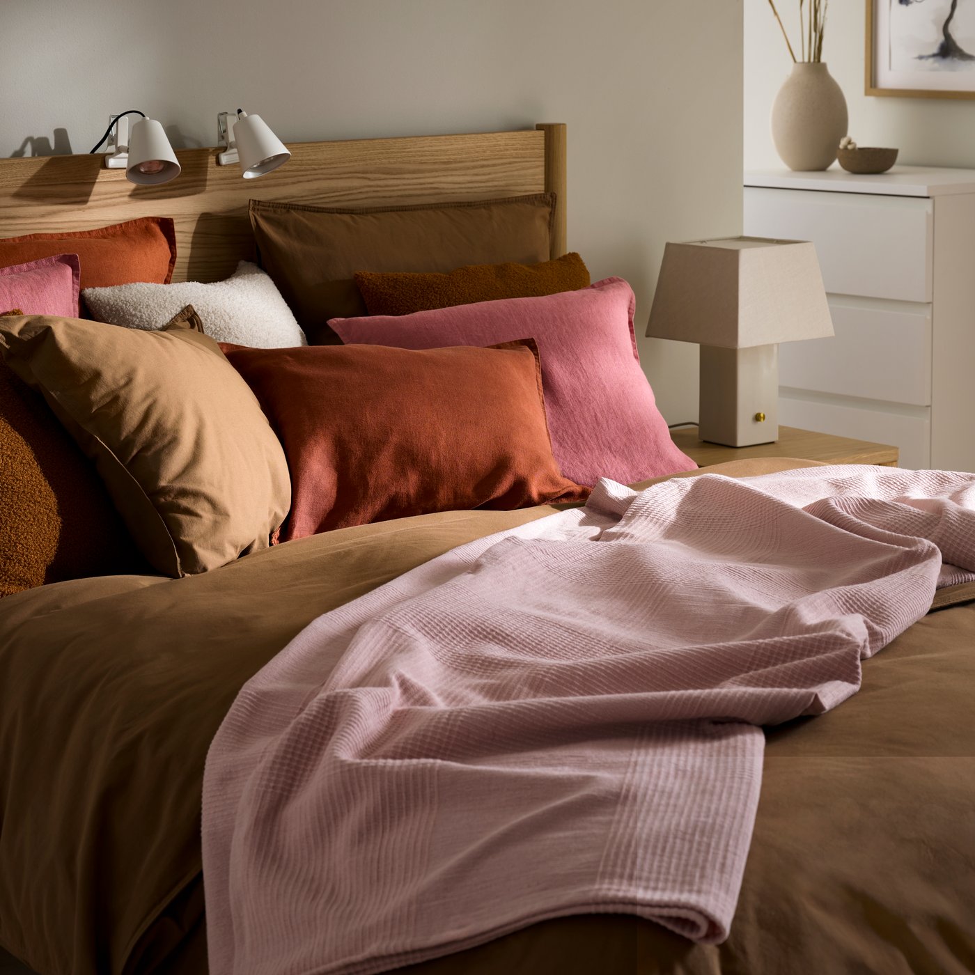 A bedroom with a bed covered in a mix of pink and brown cushions, featuring a soft pink throw, creating a cosy atmosphere.
