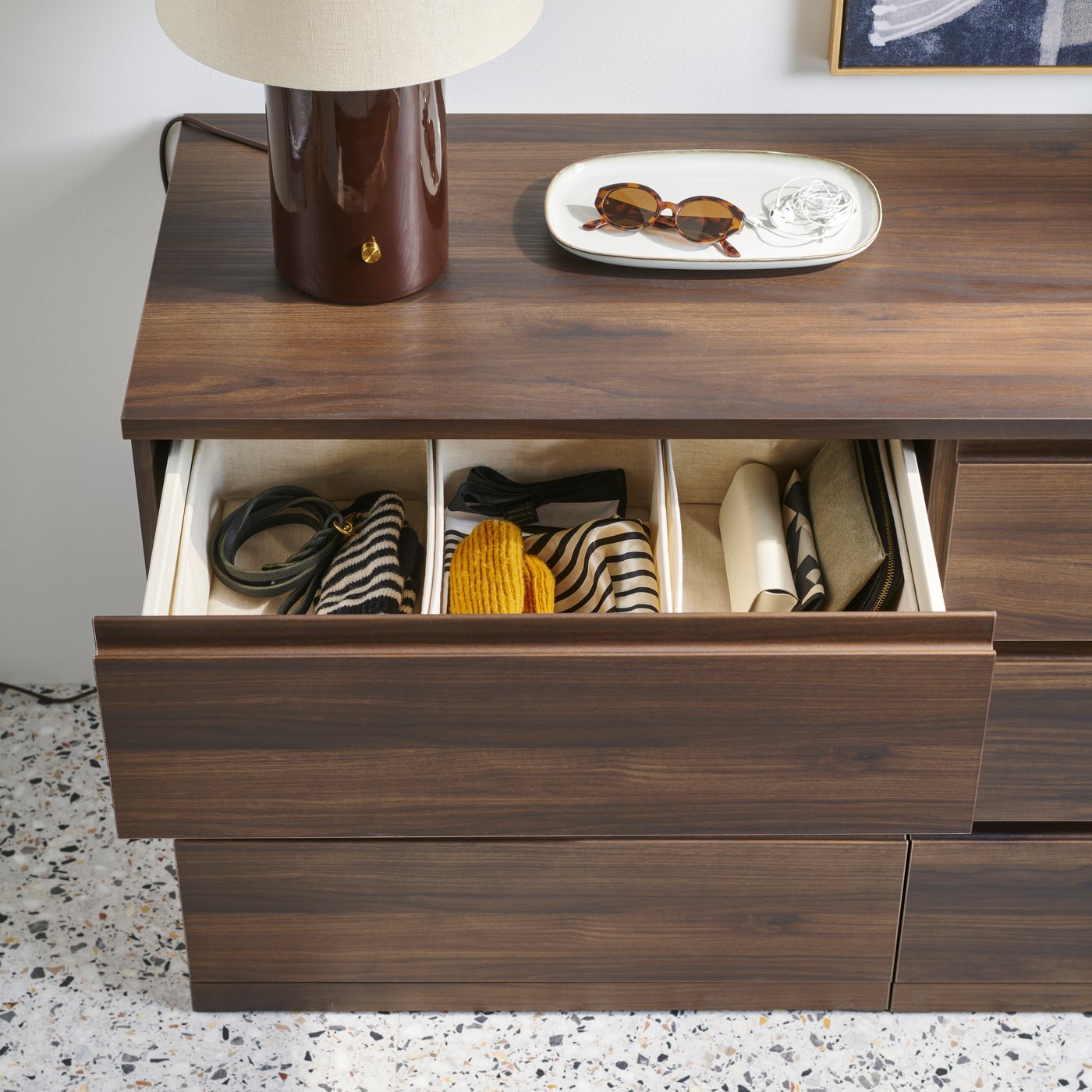 A dark brown STORKLINTA chest of 6 drawers with open drawers displaying neatly arranged items and a lamp on top.