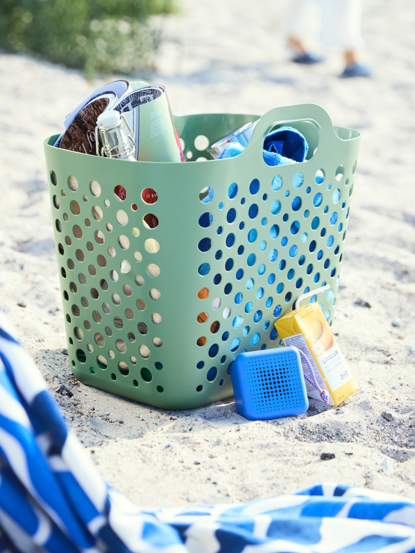 Green SLIBB laundry basket in the sand, used as a beach bag with summer items, with the blue VAPPEBY Bluetooth speaker beside.