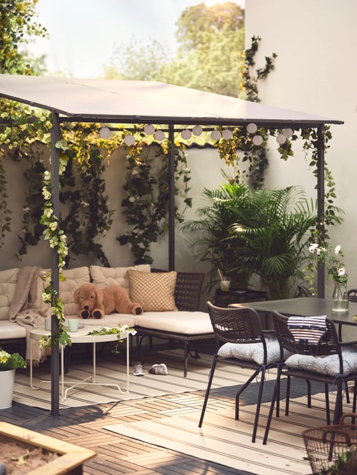 Ein GUNNÖN Pavillon auf einer Gartenterrasse mit einem Sofa und einem Couchtisch. Auf dem Sofa sind ein Stofftier, Kissen und ein Plaid zu sehen.